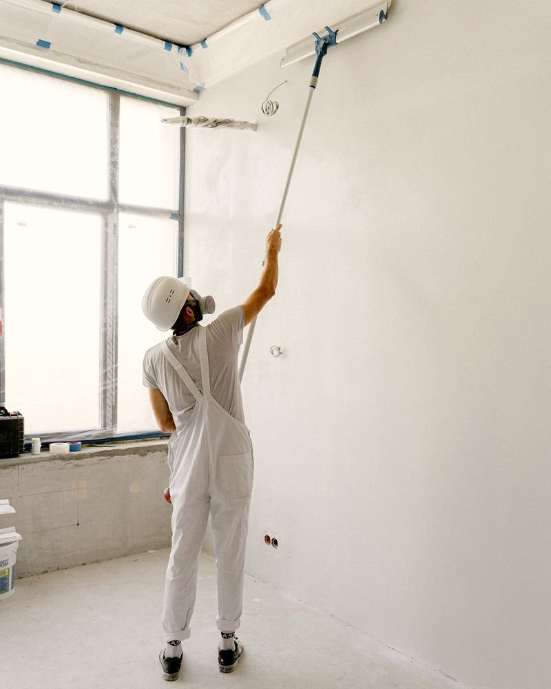 A painter in whites working on a smooth white wall, with the windows and ceiling edges sheeted and taped